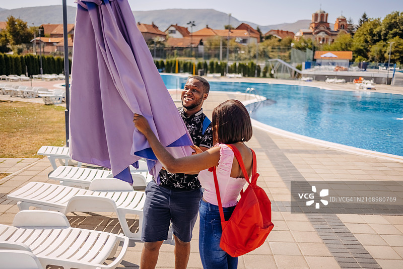 来到公共游泳池并试图打开阳伞的年轻黑人夫妇图片素材