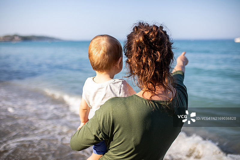 在海滩上抱着儿子的卷发年轻女子的背影图片素材