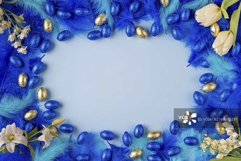 浅蓝色背景上的蓝色和绿松石色羽毛与蓝色和金色巧克力蛋、春花的边框图片素材