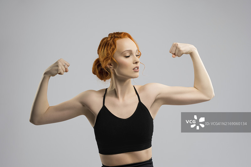 自信的年轻健美女性，姜黄色头发，黑色运动装，在灰色背景下展示肱二头肌图片素材