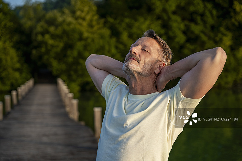 在湖边闭着眼睛，双手放在脑后的疲惫老年男人图片素材