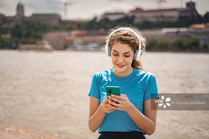 在阳光明媚的日子里，在布达佩斯多瑙河边用智能手机欣赏音乐的年轻女人图片素材
