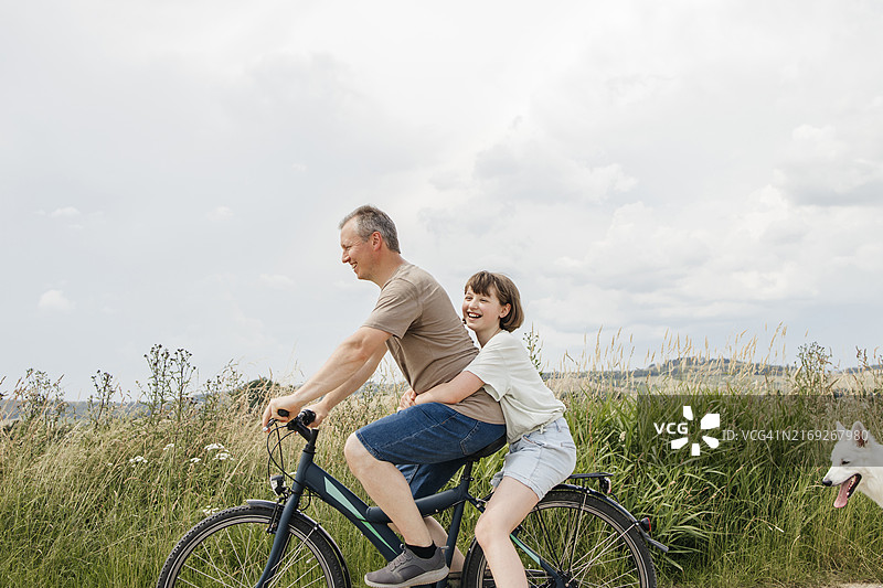 快乐的女孩坐在父亲的自行车后座，在田野附近图片素材