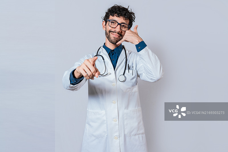微笑的医生做出打电话的手势并指向相机。做出打电话手势的年轻医生图片素材