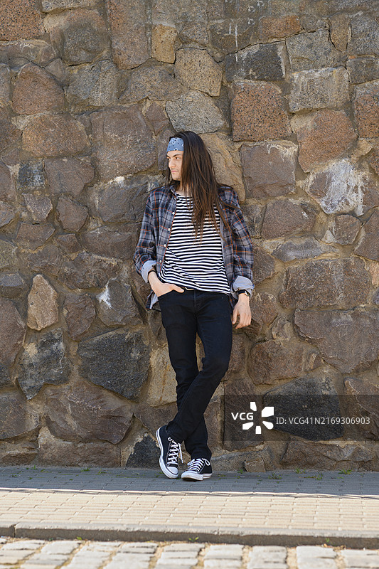 穿着格子衬衫、条纹背心和牛仔裤的年轻长发男子靠在石墙上图片素材