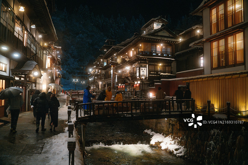 冬季，日本山形县尾花泽市的银山温泉被白雪覆盖，木制建筑和灯笼照亮的街道两侧是银山河图片素材