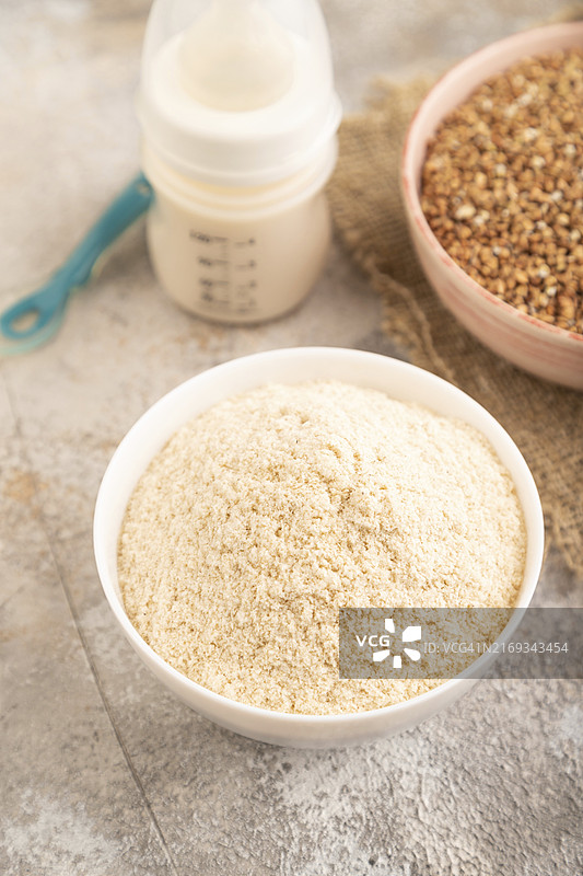 奶粉和荞麦婴儿食品混合物、婴儿配方奶粉、奶嘴、奶瓶、勺子在棕色混凝土背景和亚麻纺织品上。侧视图，特写，选择性焦点，人工喂养概念图片素材