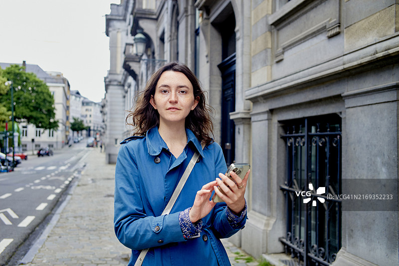 穿着蓝绿色外套的女人在阳光明媚的日子里走在迷人的欧洲城市街道上，与她的智能手机互动图片素材