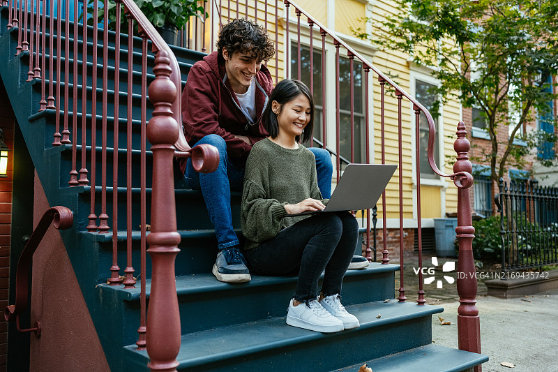 一对年轻情侣坐在都市建筑的台阶上，一起使用笔记本电脑，面带微笑。图片素材