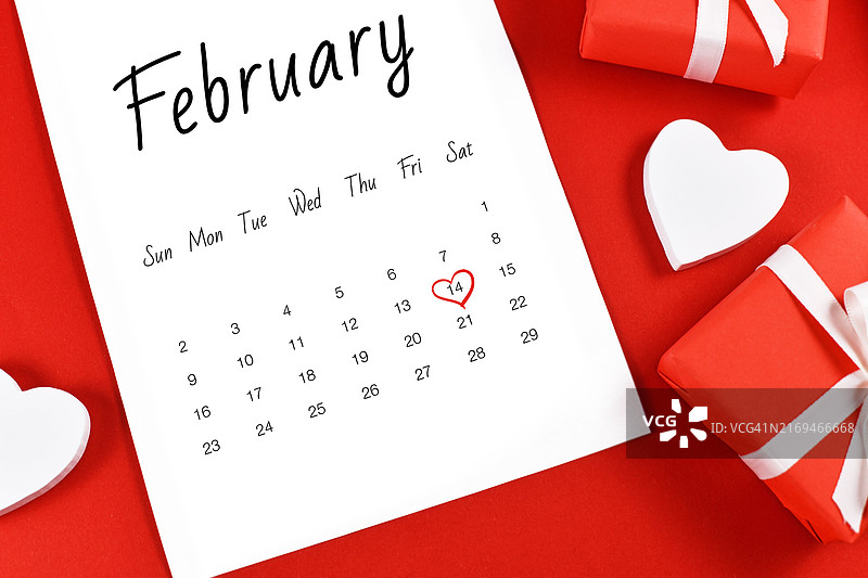 二月日历页特写，2月14日情人节用红色心形标记，周围是礼品盒和红色背景上的心图片素材