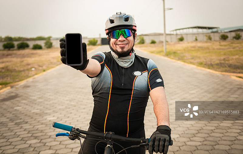穿着运动服展示手机屏幕的自行车手。在户外展示手机屏幕的男性自行车手。展示手机广告的微笑自行车手图片素材