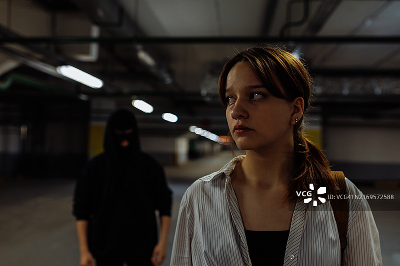 在地下停车库，一位焦虑的年轻女子看着镜头，身后站着一个戴面具的可怕男人图片素材