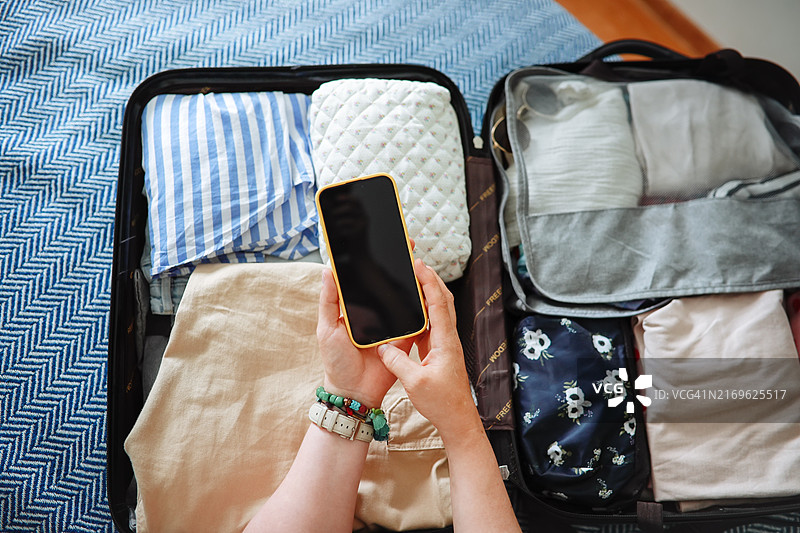 旅行App使用：大码女性手持黑屏智能手机，背景为打包好的行李箱图片素材