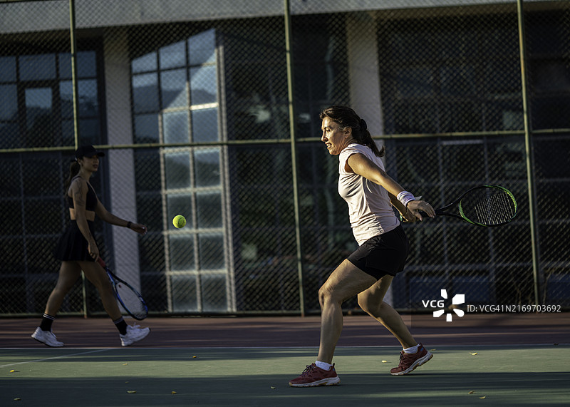 中年女网球运动员正在正手击球，练习用球在空中，另一位女网球运动员在球场上图片素材