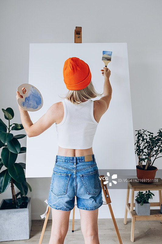 工作中的艺术家：一位戴着亮橙色无檐小便帽和休闲服装的年轻女子拿着画笔和调色板，在现代简约工作室的大画布上绘画。创造力和艺术表达图片素材