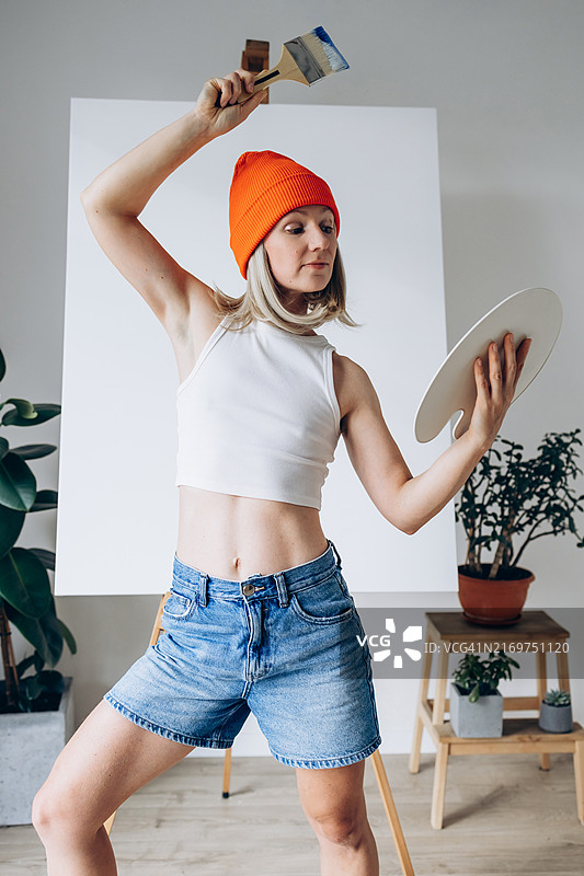 戴着橙色无檐小便帽、身穿休闲夏装的女人，手持画笔和调色板，在现代工作室中展现艺术表现的活力与激情图片素材