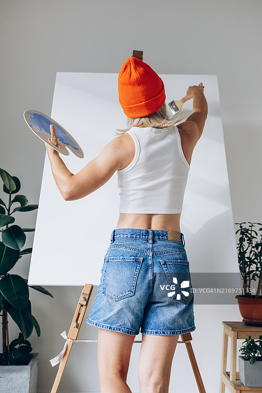 工作中的艺术家：一位戴着亮橙色无檐小便帽和休闲服装的年轻女子拿着画笔和调色板，在现代简约工作室的大画布上绘画。创造力和艺术表达图片素材