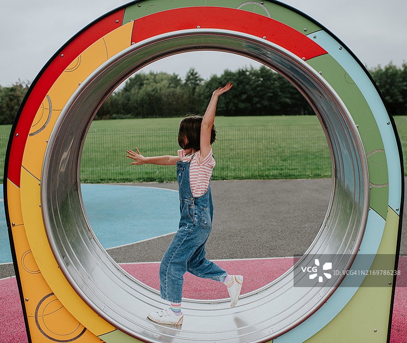 在多云的一天，年轻女孩在绿草茵茵的公园里，快乐地在一个色彩缤纷的操场仓鼠轮状结构里玩耍图片素材