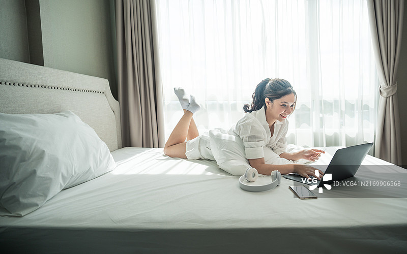 快乐微笑的亚洲女人穿着白色睡衣躺在舒适的床上，用信用卡在笔记本电脑上进行网上购物，早上在卧室里成功支付图片素材