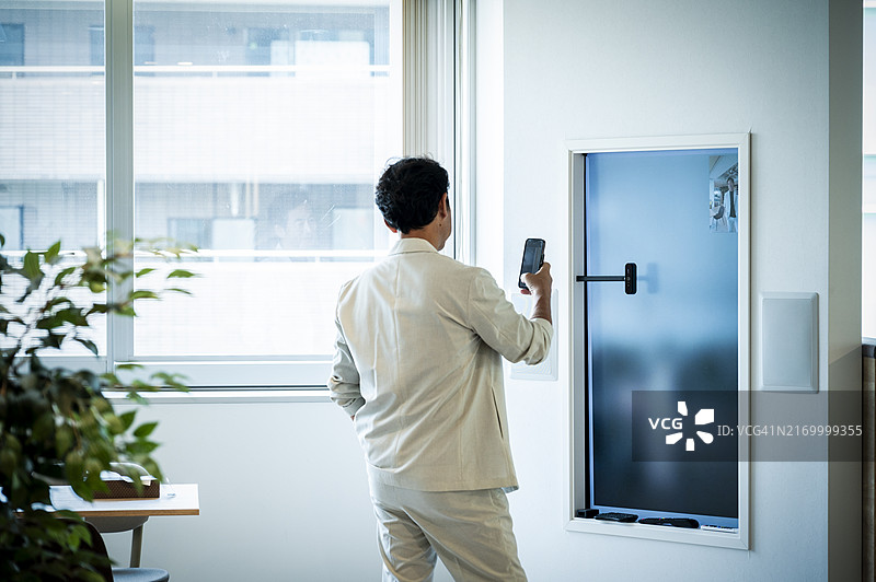 一位身穿西装的男士站在办公室的大屏幕前，正在进行远程沟通。他手中拿着一部智能手机。图片素材
