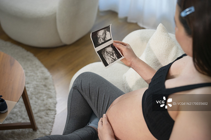 在客厅里看着并轻轻触摸隆起的腹部的年轻漂亮亚洲孕妇，准妈妈，等待新生儿，爱和亲情图片素材