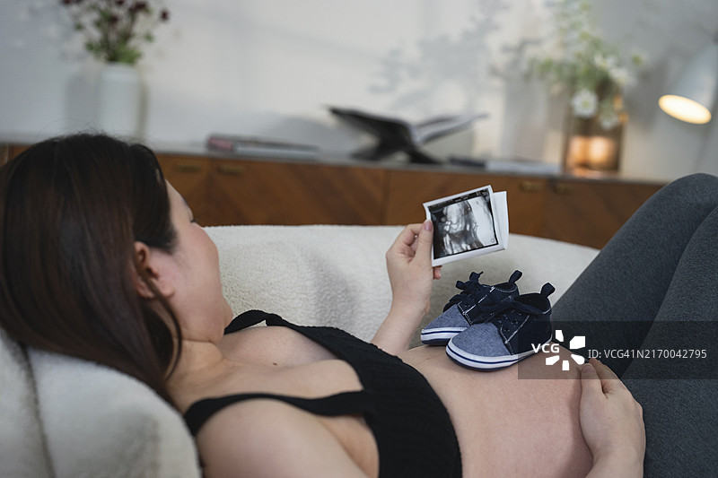 在客厅里看着并轻轻触摸隆起的腹部的年轻漂亮亚洲孕妇，准妈妈，等待新生儿，爱和亲情图片素材