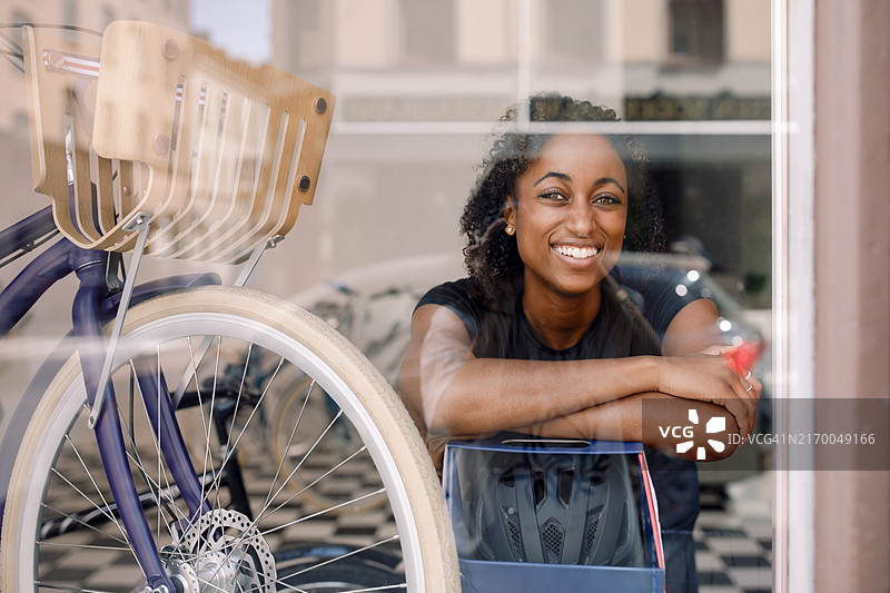 透过自行车商店的窗户看到的，倚靠在自行车旁边的快乐女店主肖像图片素材