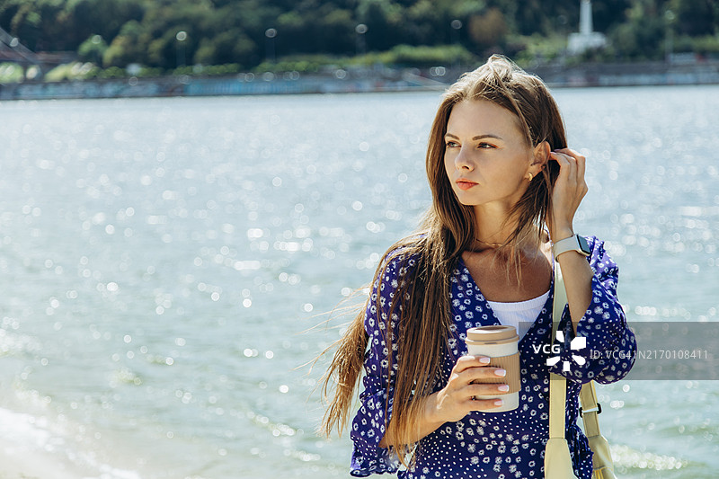 年轻女人在明亮的城市里，在河边看着远方，手里拿着一次性杯子图片素材