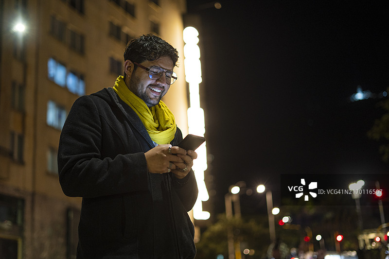 千禧一代的男人走在城市街道上，同时通过智能手机与朋友交谈图片素材