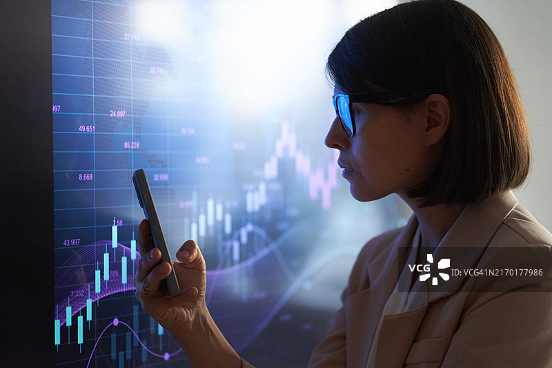 在投资图表背景下的使用智能手机的商务女性的侧面照图片素材