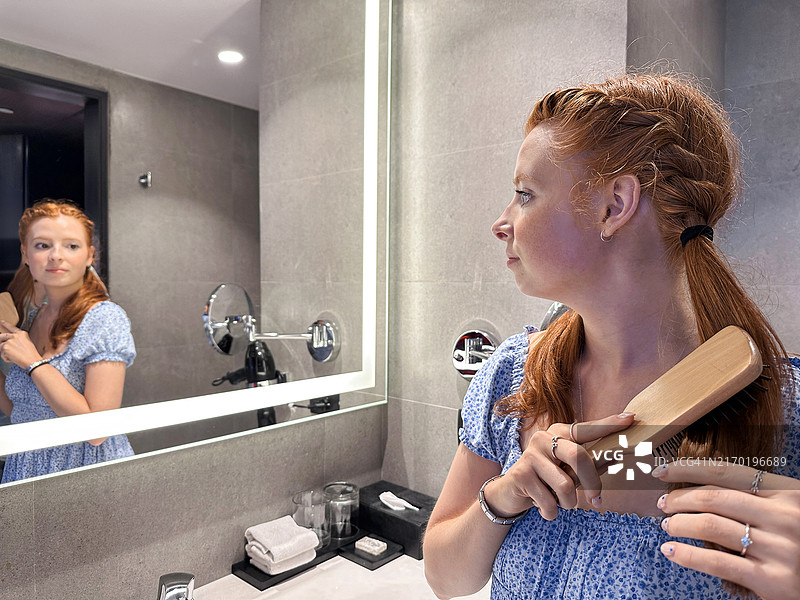 迷人的红发女子站在白色浴室水槽前，穿着太阳裙，在明亮的镜子中反射，使用发刷，梳理头发，侧面视角，自我护理和仪容概念特写图片素材