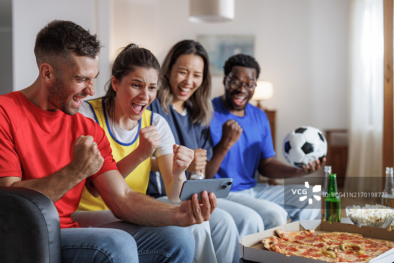 朋友们坐在沙发上，喝着啤酒，吃着披萨，专注地看着智能手机上的足球比赛图片素材