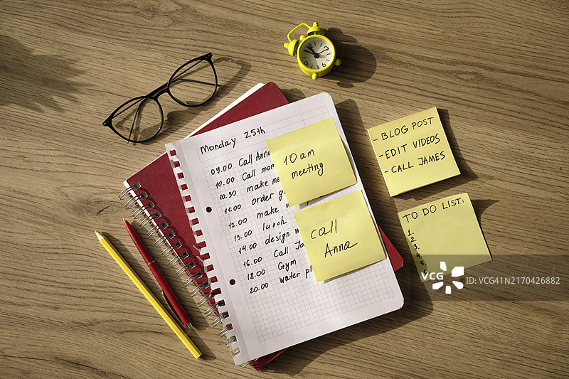 带有任务的粘性黄色便签，工作桌上的待办事项记事本。办公用品、眼镜、铅笔、时钟放在木质表面上。图片素材