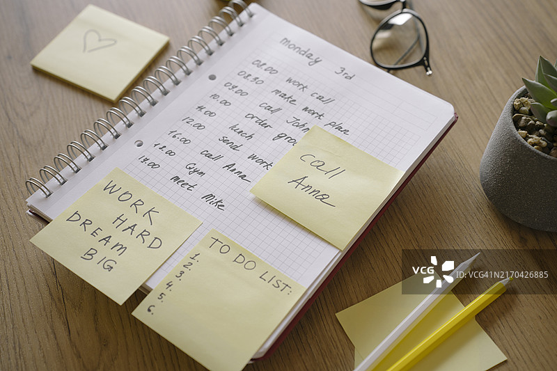 带有任务的粘性黄色便签，工作桌上的待办事项记事本。办公用品、眼镜、铅笔、时钟放在木质表面上。图片素材