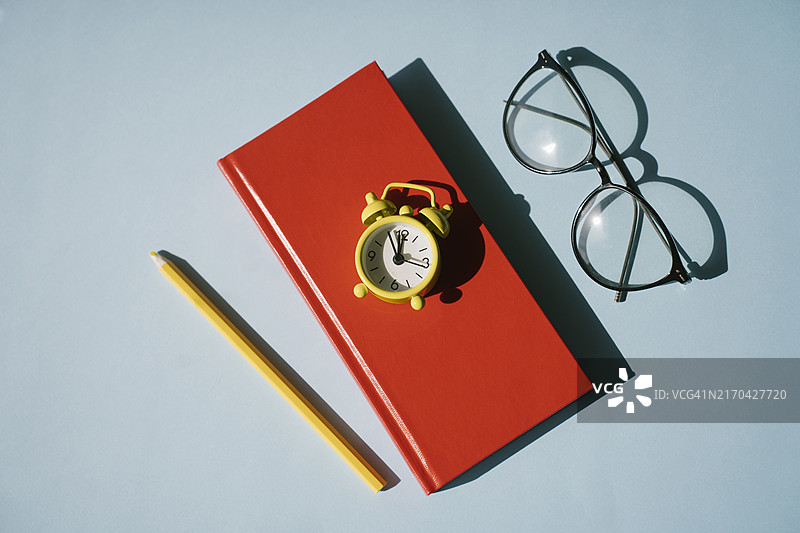 蓝色背景上的红色笔记本、黄色闹钟、铅笔和眼镜，阳光直射图片素材