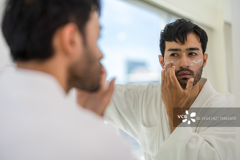 亚洲男人在浴室镜子前涂抹面霜，注重健康保湿的护肤程序，现代生活中的自我护理和仪容图片素材