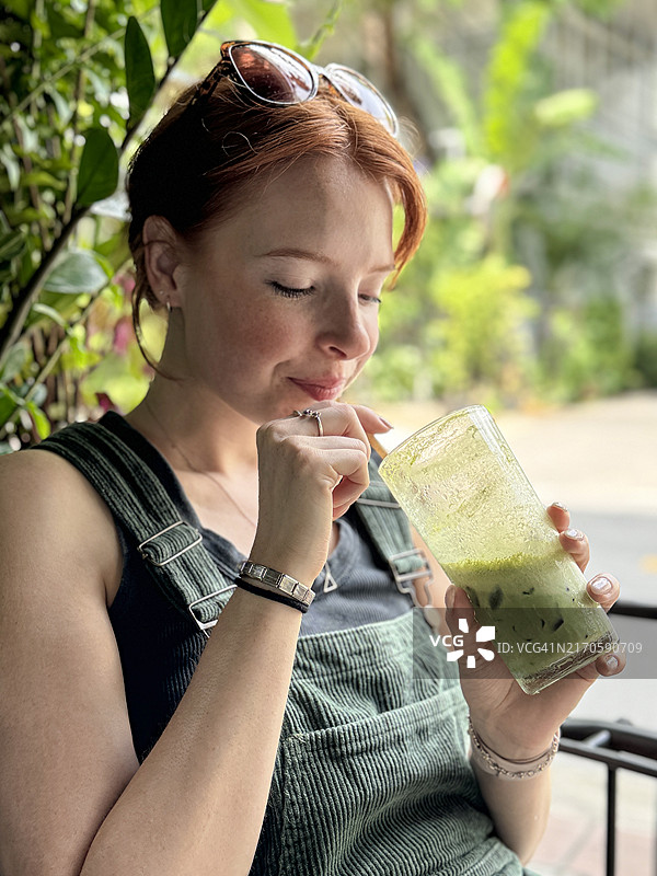 迷人的红发女子拿着冰抹茶奶昔，坐在户外用餐区图片素材