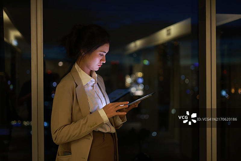 年轻女商人在金融区使用平板电脑，背景是夜间灯火通明的公司摩天大楼图片素材