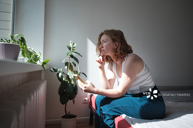 悲伤沮丧的女人看着智能手机，坐在床上用手机阅读坏消息，在社交媒体上滚动浏览，感到难过。小工具与精神健康图片素材