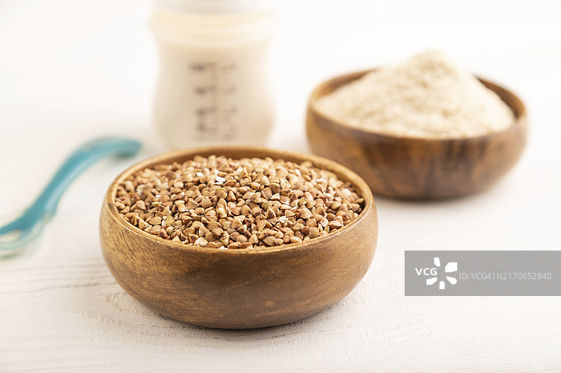 奶粉和荞麦婴儿食品混合物、婴儿配方奶粉、奶嘴、奶瓶、勺子在白色木质背景上。侧视图，特写，选择性焦点，人工喂养概念图片素材