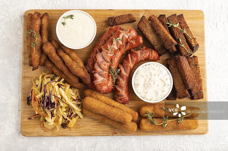 一组小吃：香肠、炸鸡块、奶酪条、吐司、卷心菜沙拉，灰色水泥背景的砧板上。俯视图，平铺，特写图片素材