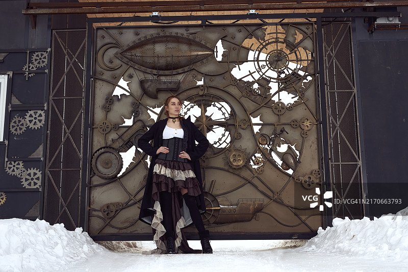 年轻女子穿着复古蒸汽朋克服装，靠在旧金属门上图片素材