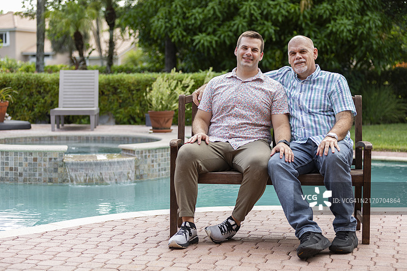 父亲和年轻的成年儿子坐在房子后院游泳池边的木凳上图片素材