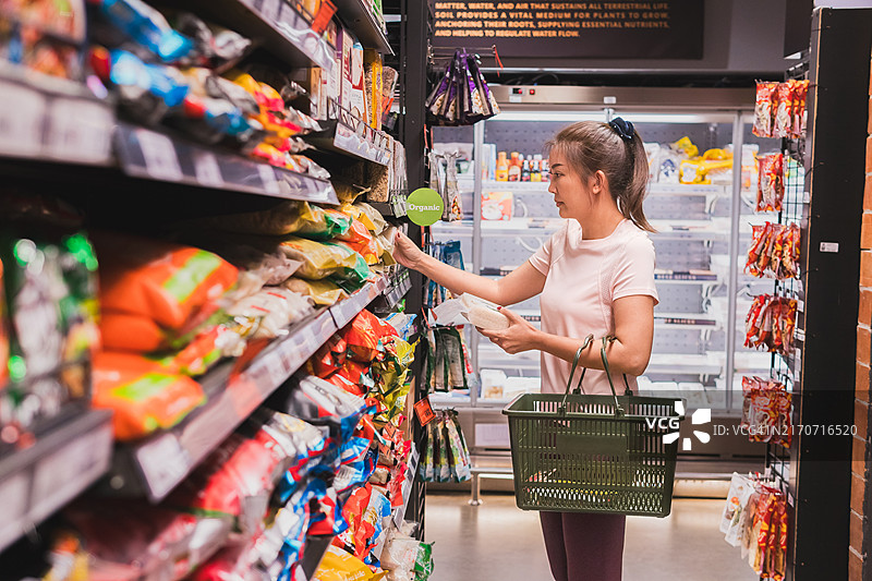 亚洲女人在超市拿着篮子，比较货架上不同的米，关注健康营养产品的选择，日常购物中注重消费者行为图片素材