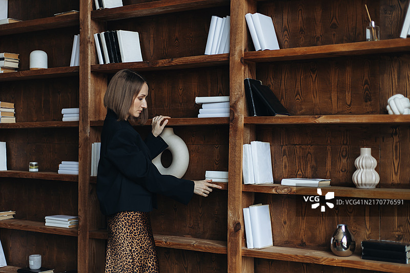 穿着夹克和豹纹裙的年轻女人触摸着摆满书籍的木制书架图片素材