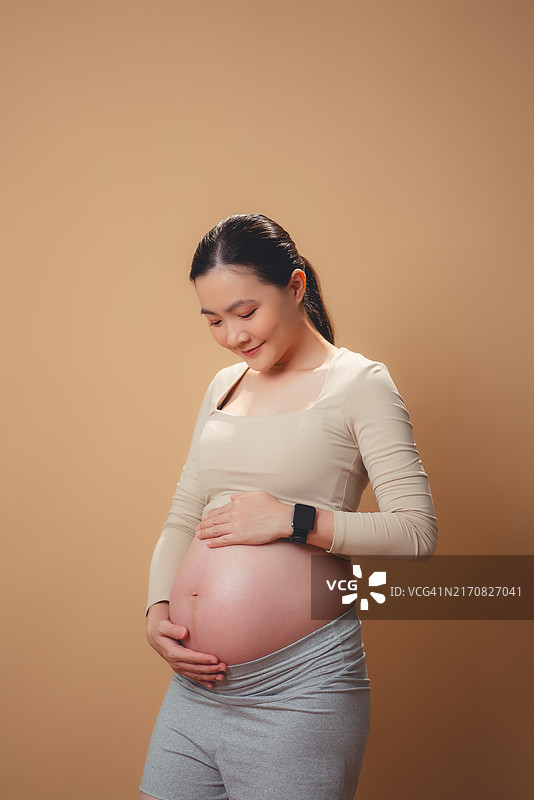 亚洲孕妇幸福地笑着看着镜头，隔离在米色背景上图片素材