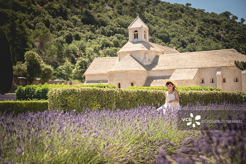 小女孩戴着草帽，穿着白色连衣裙在法国普罗旺斯沃克吕兹省戈尔德村附近的薰衣草田中奔跑图片素材