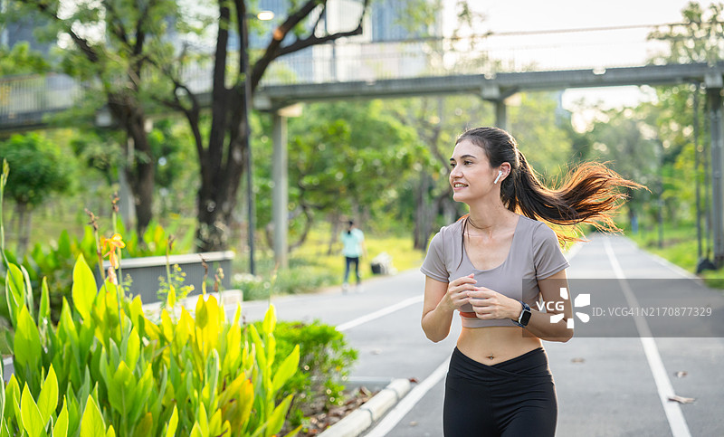 年轻女子在公园小径上慢跑，周围绿树环绕，背景是人行天桥，她看起来充满活力，非常健康。图片素材