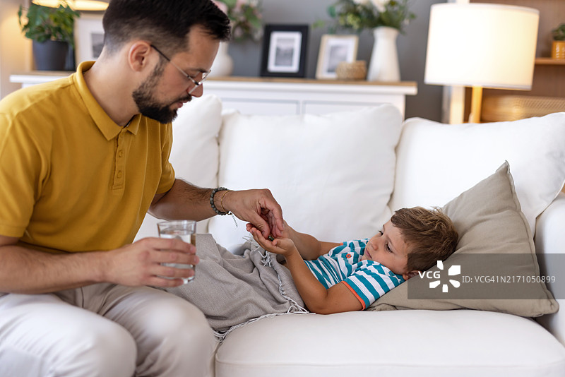 父亲在白色沙发上给生病的孩子喂水图片素材