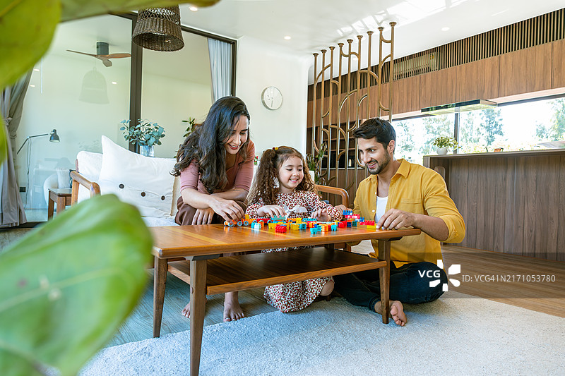 在一个现代简约的家居环境中，父母积极参与活动，微笑着帮助女儿搭建积木，突显了他们之间的亲密关系和团结。图片素材